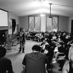 Speaker addressing a large, attentive audience in a dimly lit room.