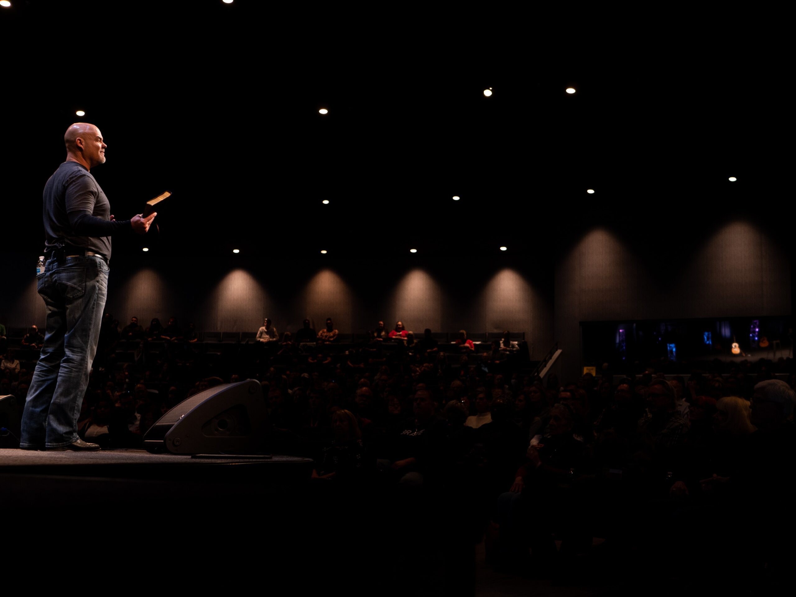 Man speaking on stage holding a book in front of a seated audience in a dark room.